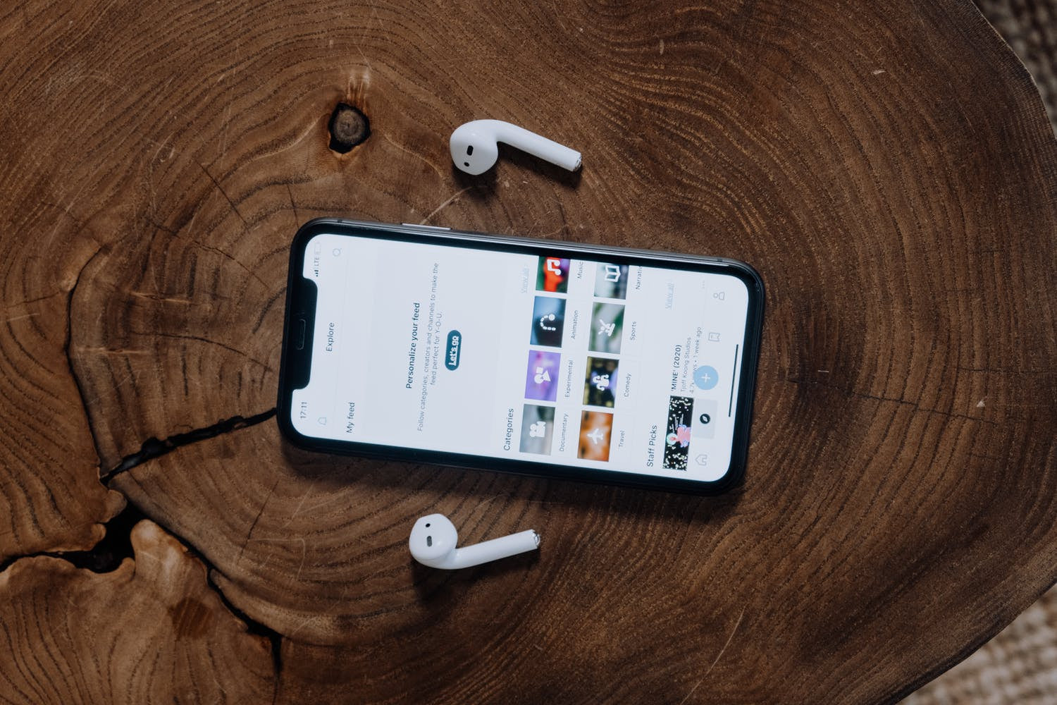 Smartphone in a wooden case on a wooden table with wireless earbuds, showcasing a playlist on the screen.