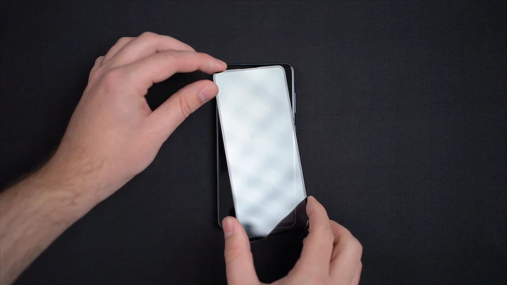 Person applying tempered glass screen protector to smartphone on a dark background.