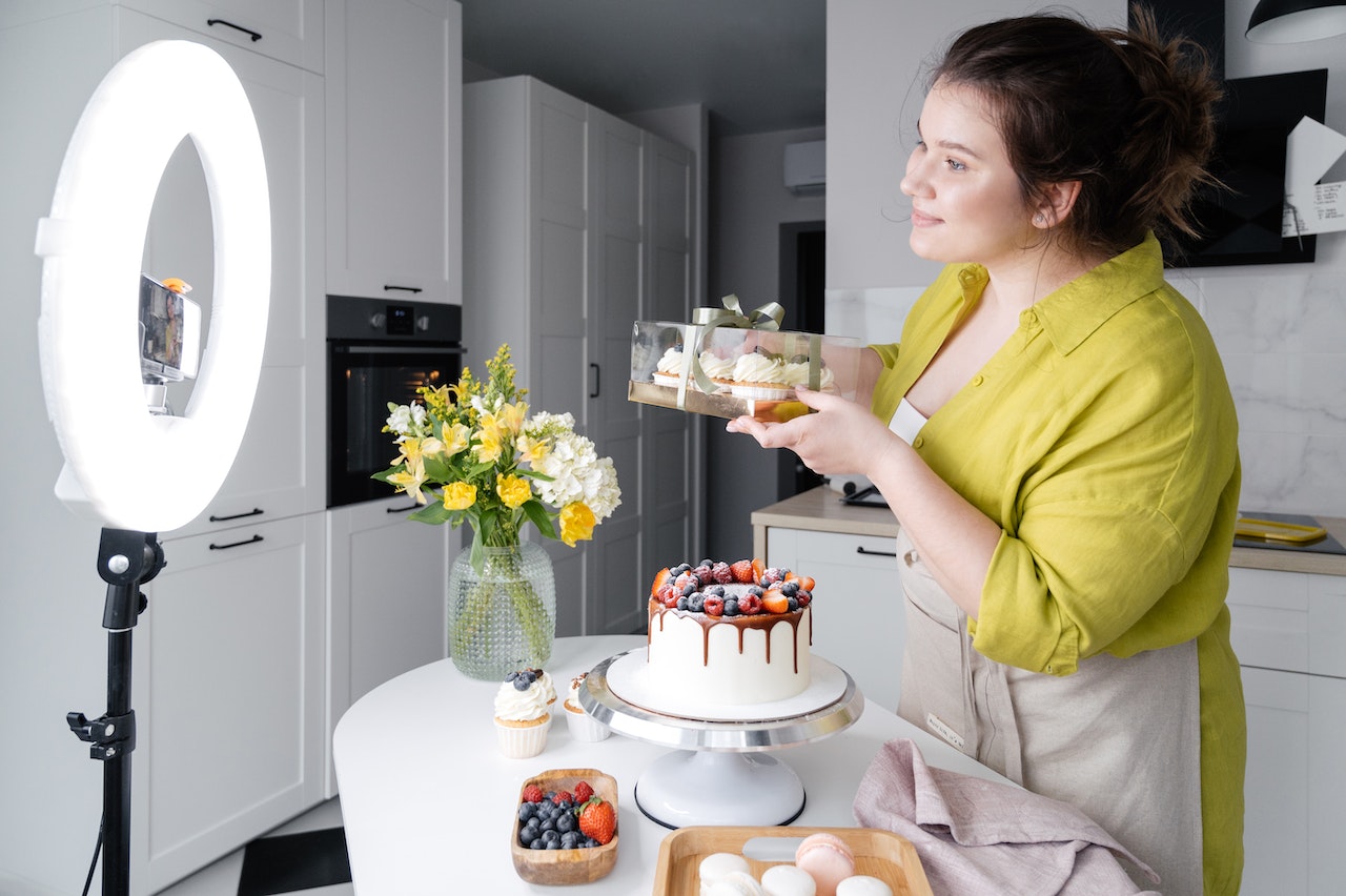 Frau backt Kuchen mit LED-Ringlicht im Hintergrund, dekoriert mit Blumen und Beeren in moderner Küche.