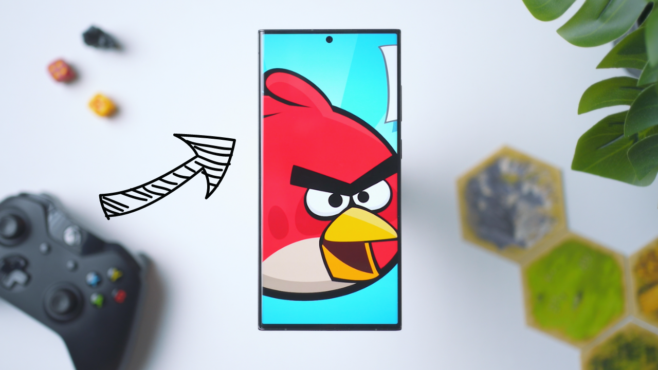 Smartphone displaying Angry Birds with gaming setup, including controller and board game, on a white surface