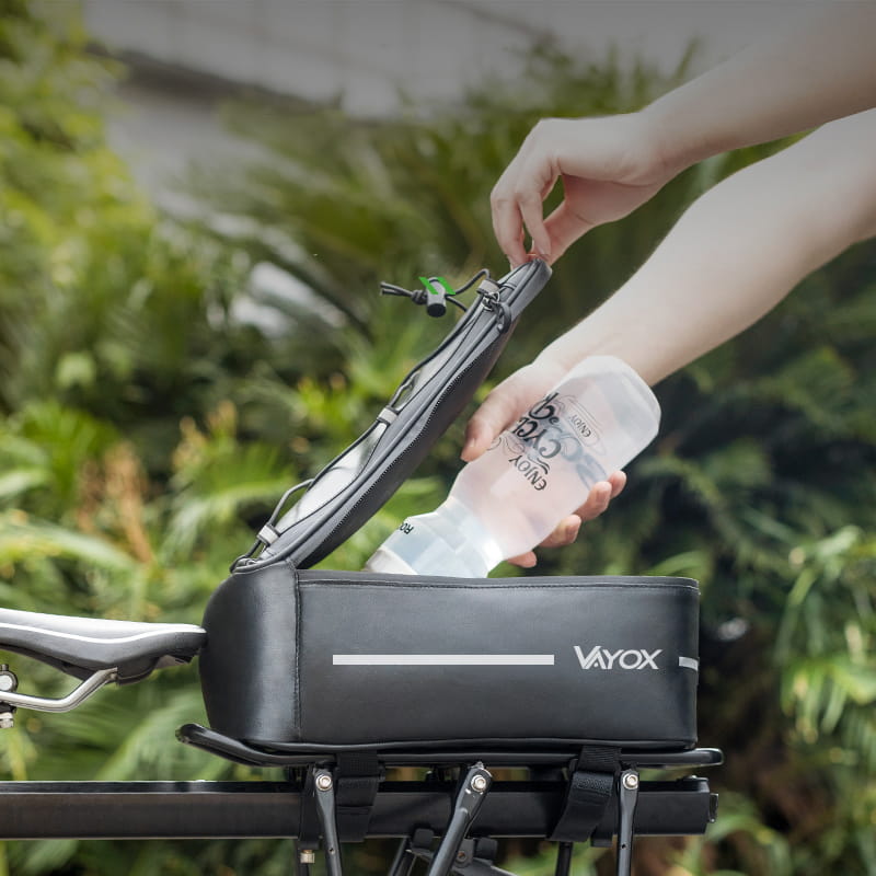 Bicycle rear rack with black Vayox VA0166 8L pannier bag, open lid, showing water bottle being placed inside, lush greenery background.