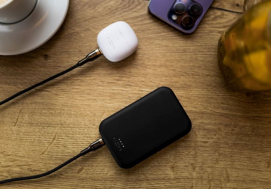 Schwarze Powerbank Fixed MagZen 6, 6000 mAh, lädt kabellos ein Gerät neben einem iPhone und AirPods auf einem Holztisch.