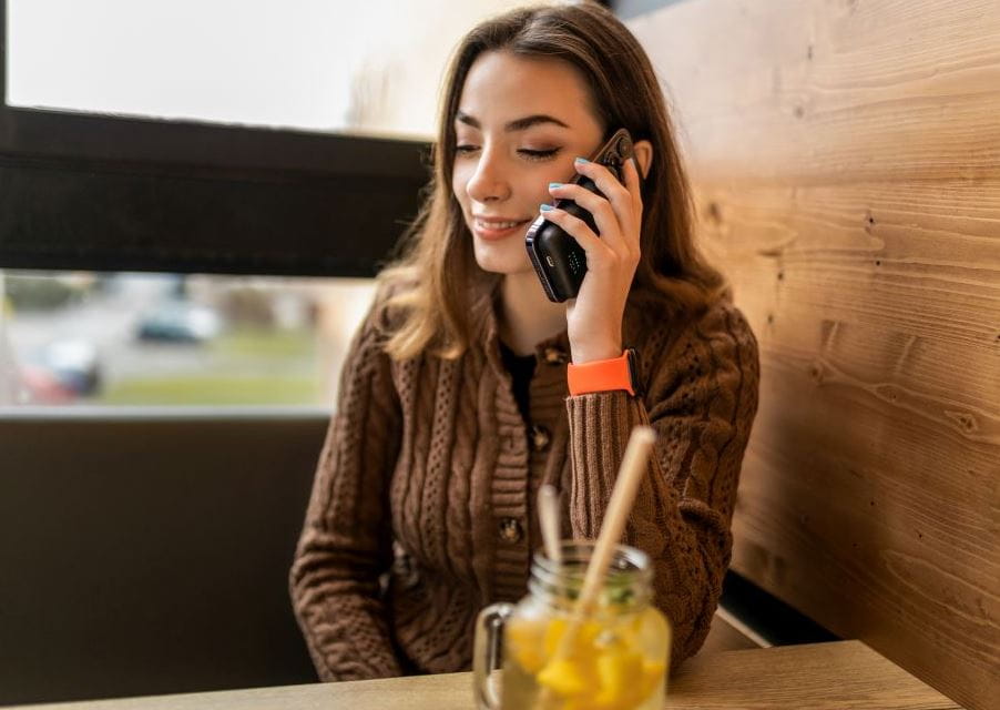 Frau spricht lächelnd in ein Smartphone im Café, trägt braune gestrickte Jacke und orangenes Armband, vor einem Glas Zitronenwasser.