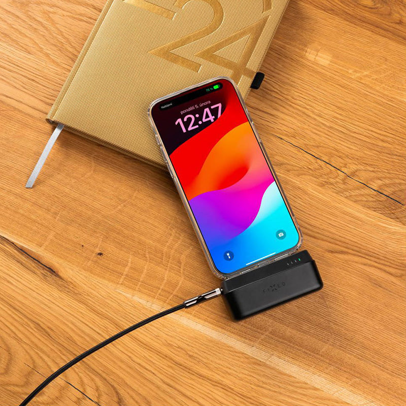 Smartphone wird mit schwarzer Powerbank Fixed Zen 5 auf Holztisch aufgeladen, neben einem goldenen Notizbuch, stromsparend und praktisch.