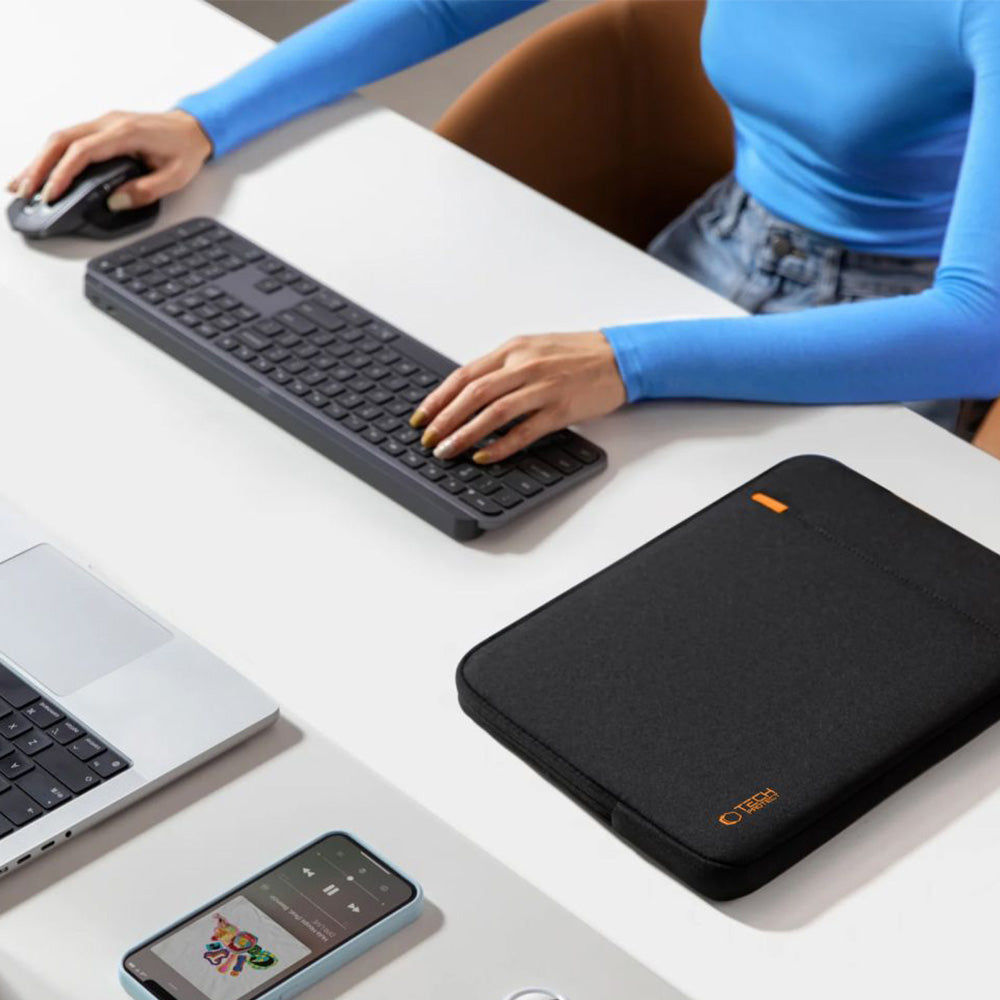 Person using a keyboard and mouse beside a black laptop sleeve on a desk.